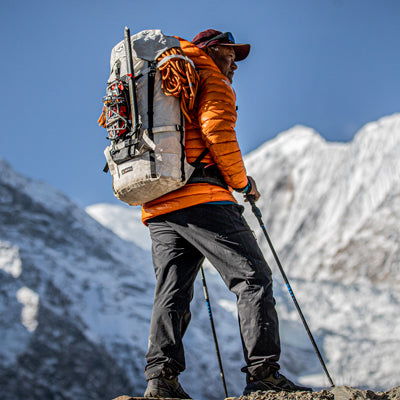 Alpine climber with a Hyperlite Mountain Gear Halka pack loaded with rope, standing on a rocky ridge with glaciated peaks in the background.