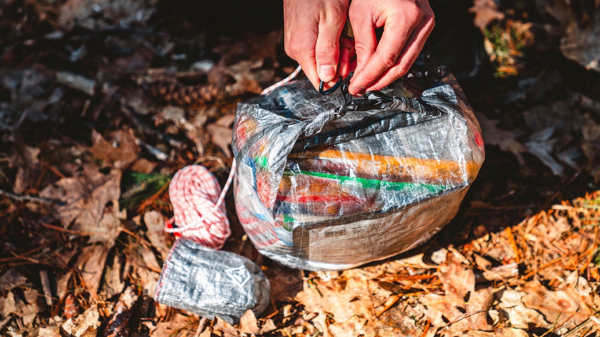 A person's hand holding a sealed plastic bag full of colorful waste, on a forest floor scattered with dry leaves.