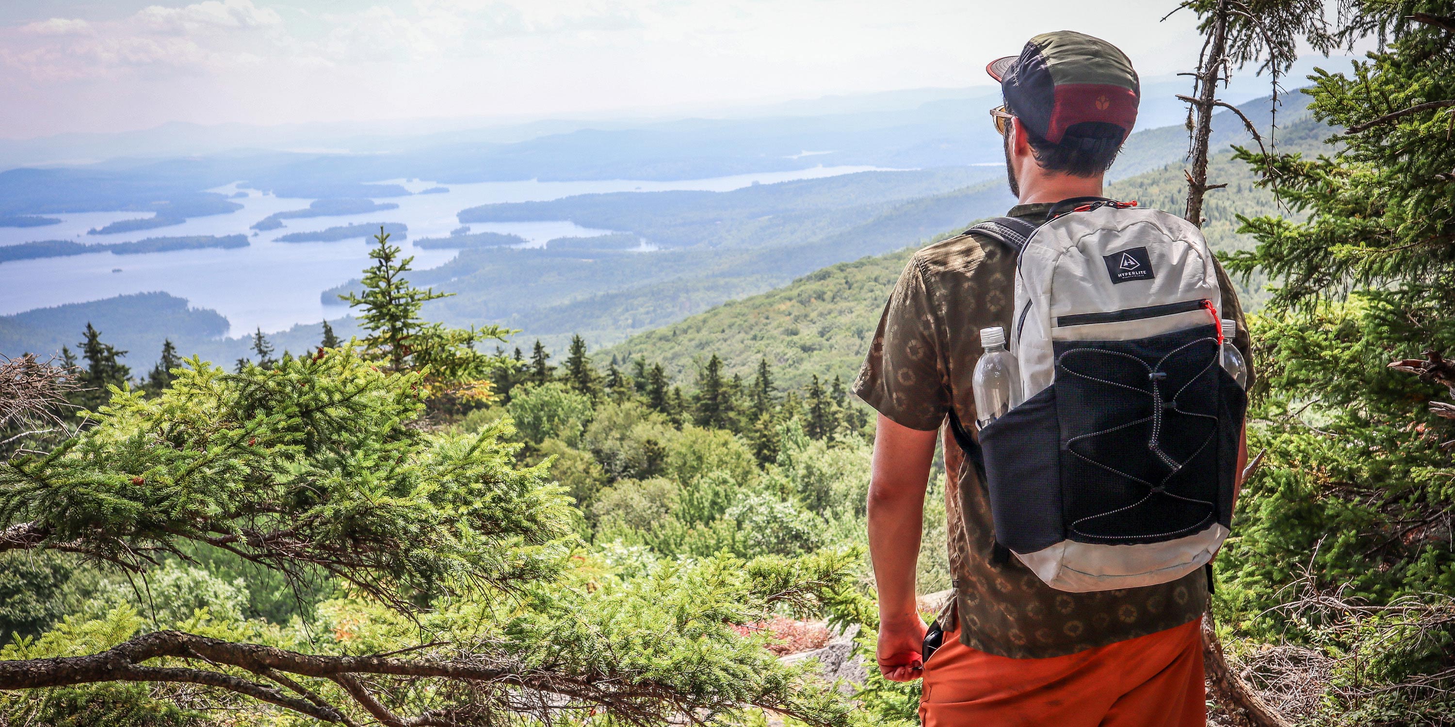 Man with a Hyperlite Mountain Gear Daybreak 22 backpack overlooking a scenic landscape with trees and water.