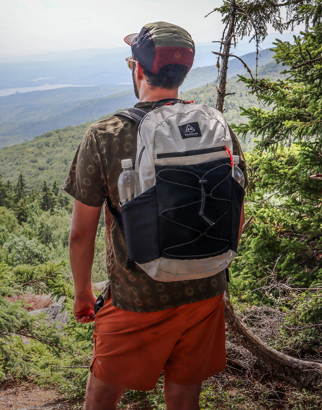 Man with a Hyperlite Mountain Gear Daybreak 22 backpack overlooking a scenic landscape with trees and water.