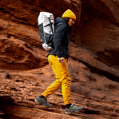 Hiker with a yellow beanie and a Hyperlite Mountain Gear Elevate 22 daypack walking alongside a steep, red rock wall in a desert canyon.