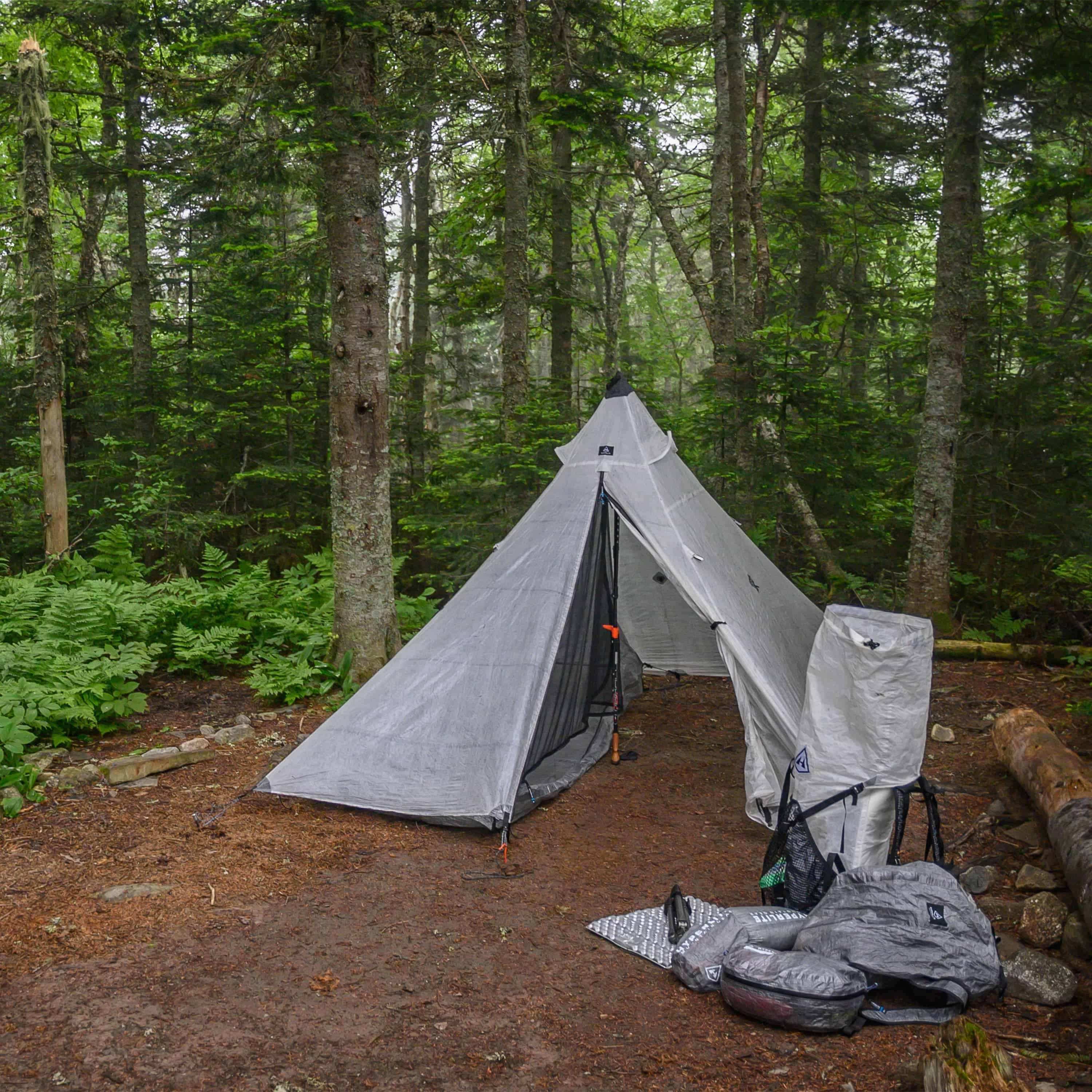 Front exterior view of the Hyperlite Mountain Gear Shelters UltaMid 2 Half Insert inside white tent in a wooded setting. Assorted Hyperlite hiking gear sits beside the outside right of the tent