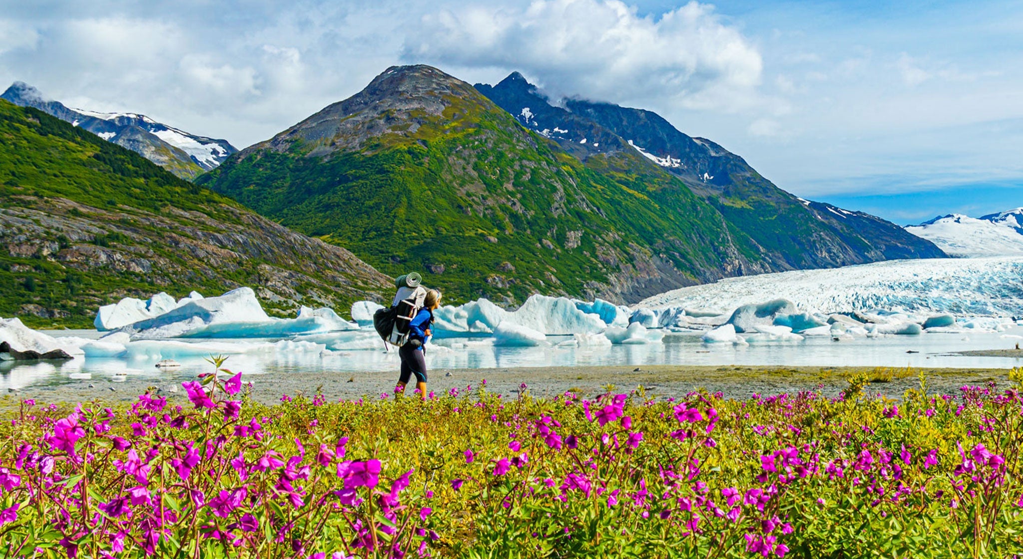 A woman is standing in front of a glacier with flowers in the background.