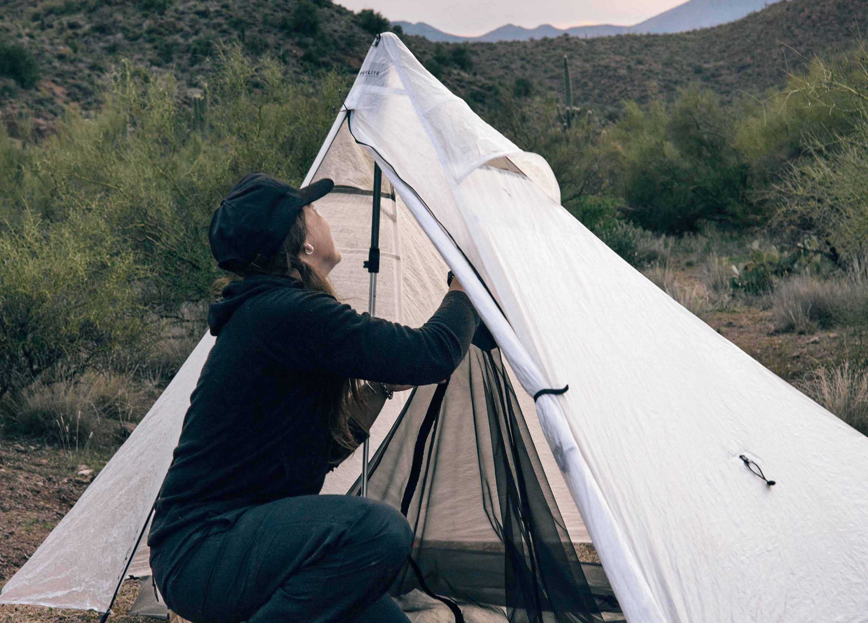 A person in a black cap and coat setting up a white tent in a desert area during dusk.