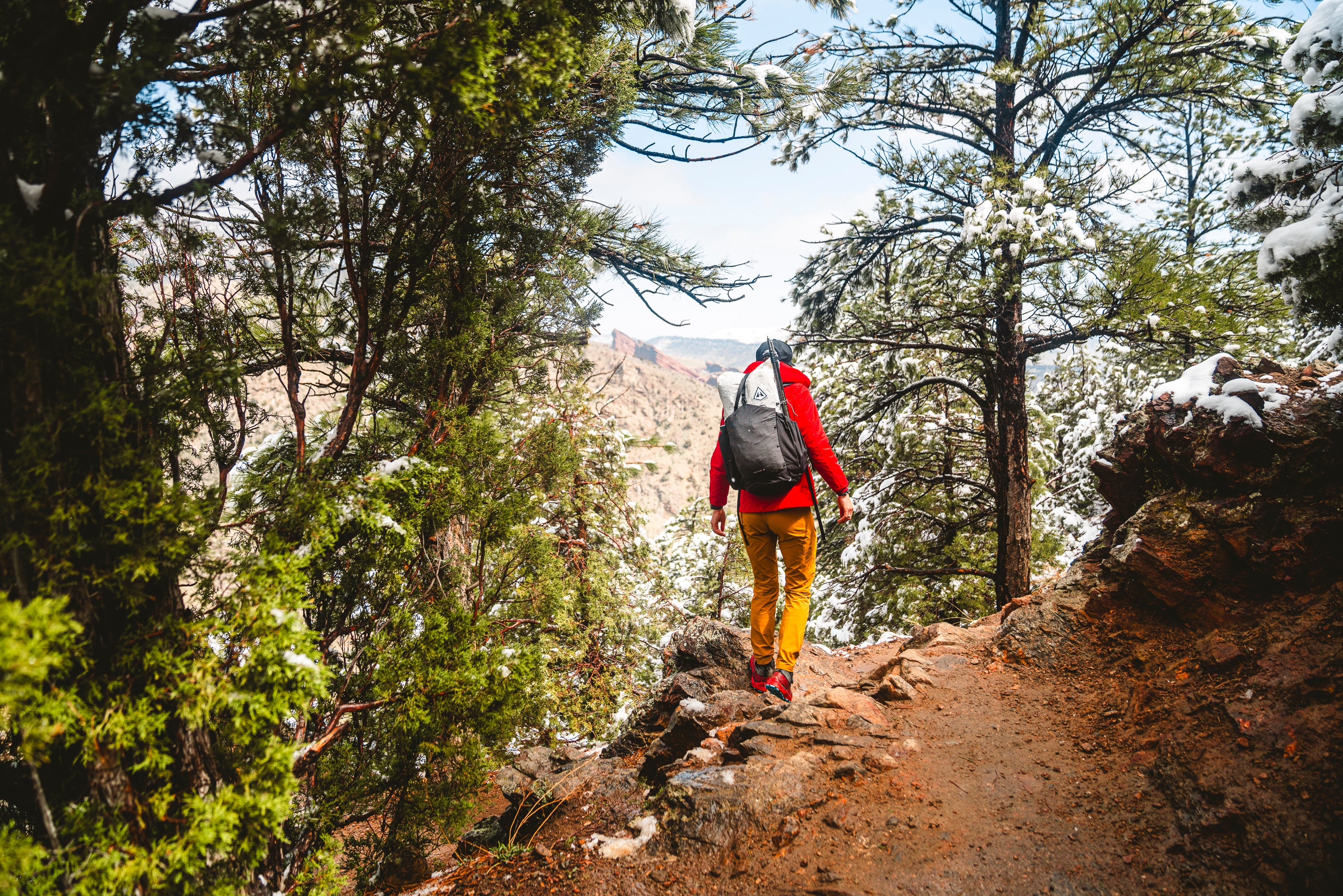 A hiker wearing a red backpack and yellow pants walking on a forest trail with patches of snow, overlooking a canyon.