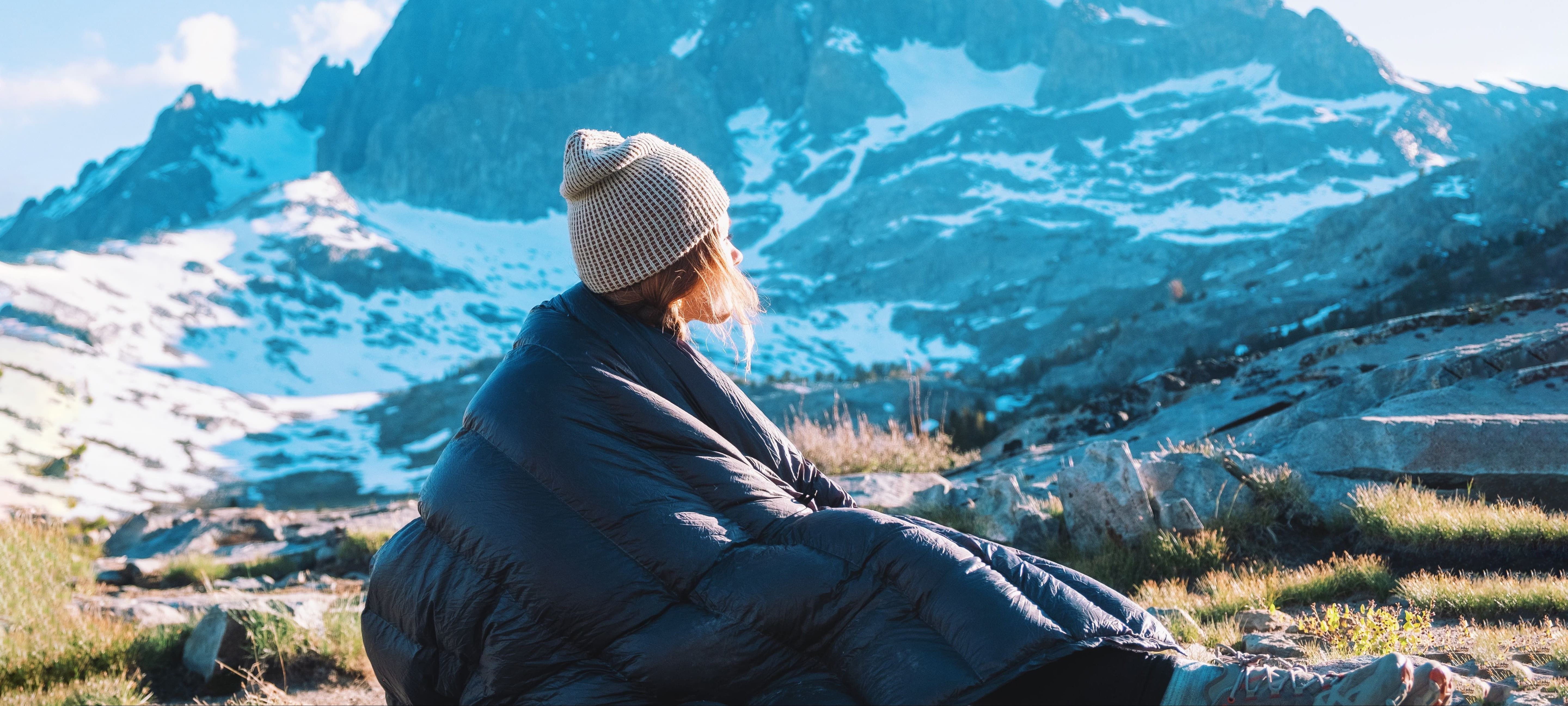 A woman sitting in a sleeping bag in front of mountains.