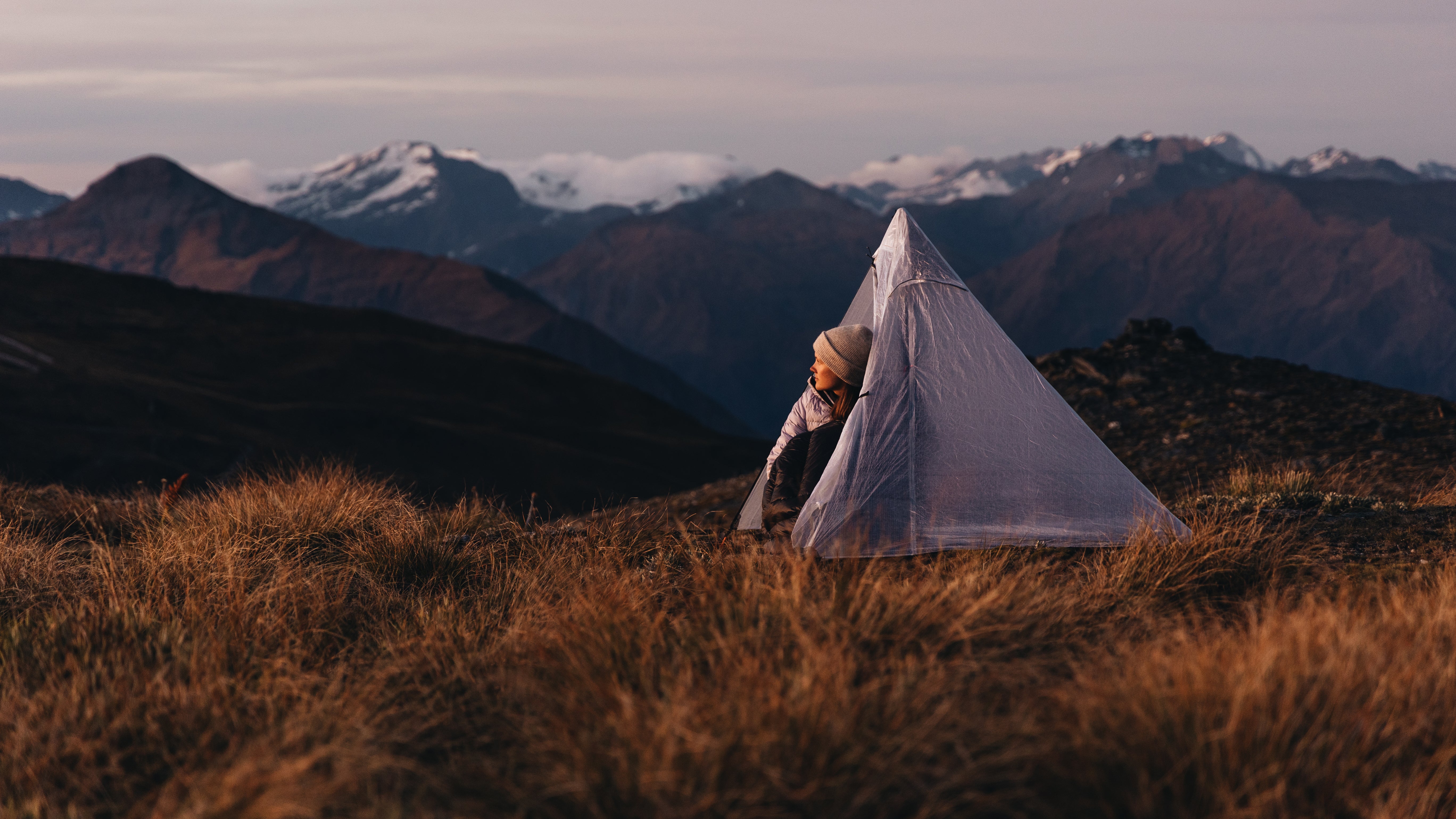 A person setting up a triangular tent on a grassy mountain ridge at dusk, with snow-capped peaks in the background.