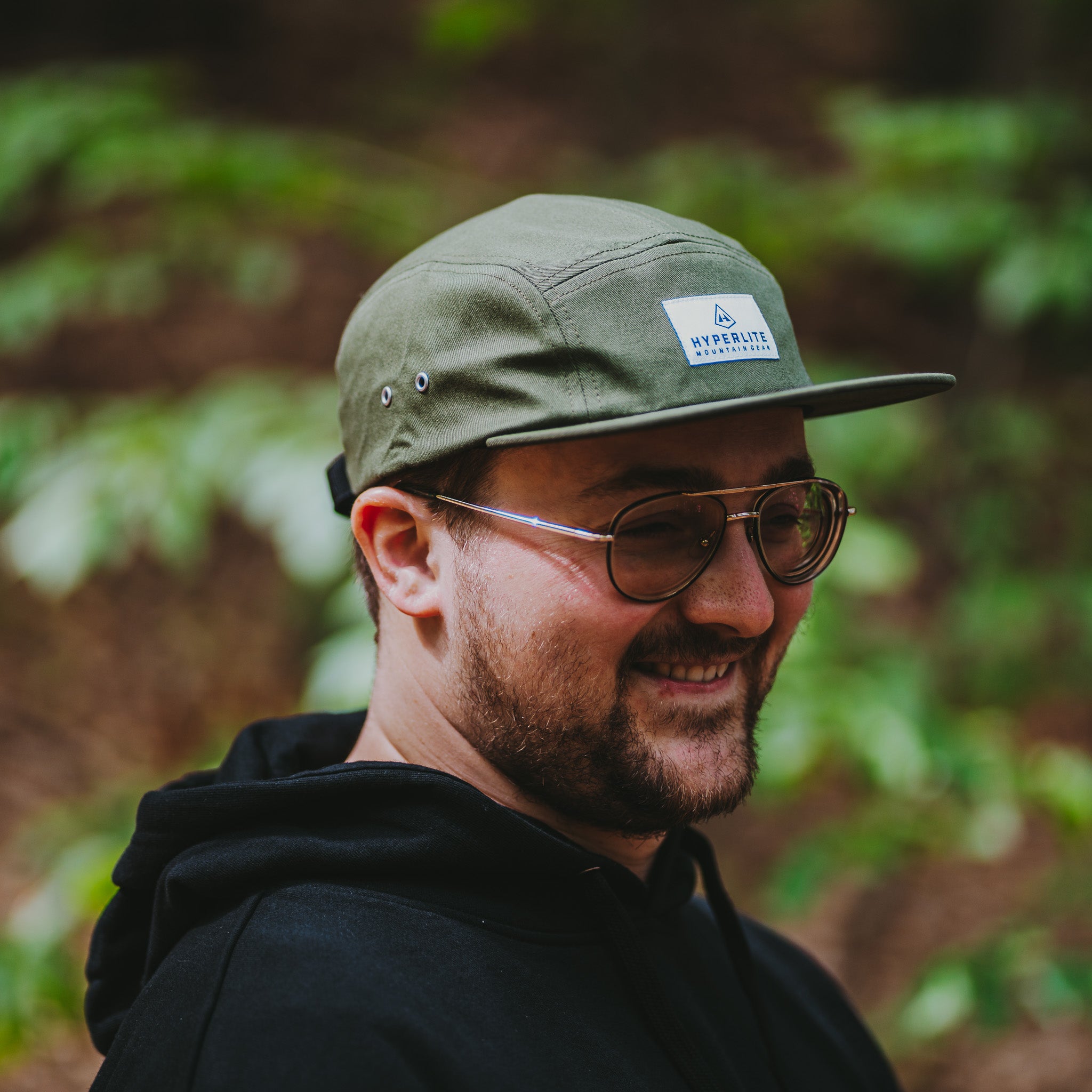 Man wearing a green cap with a Hyperlite Mountain Gear logo and sunglasses, standing in a forest.
