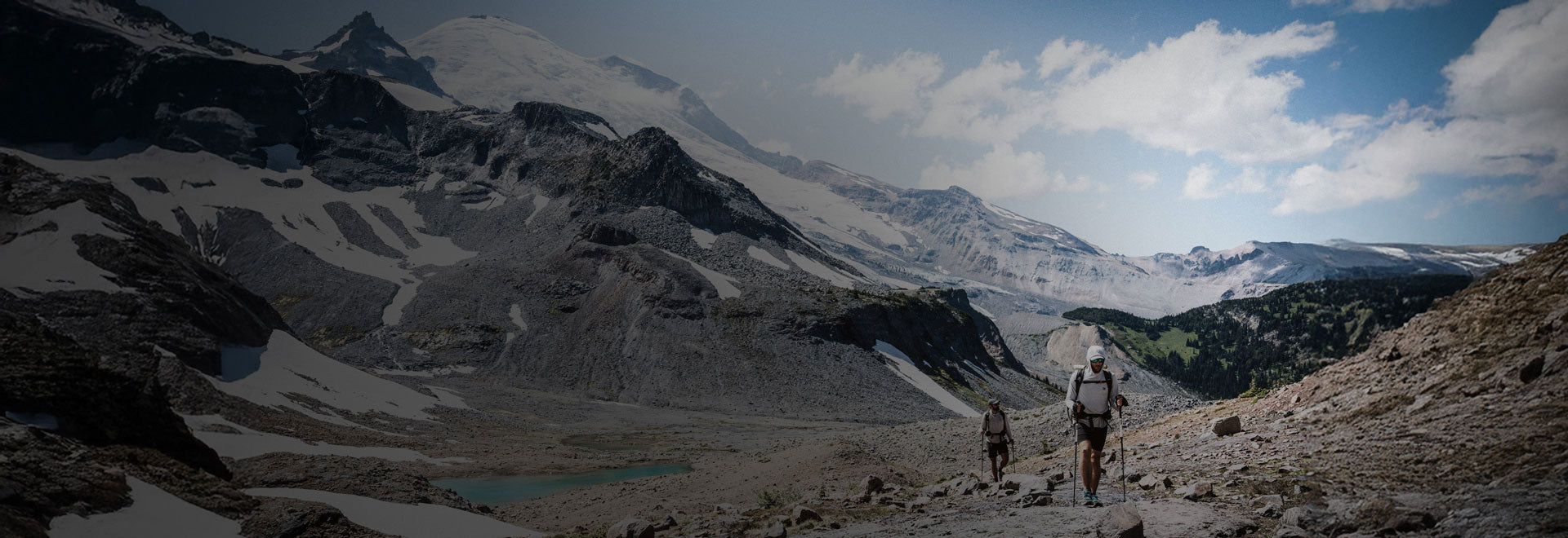 Two hikers on a mountain trail with snow-capped peaks and a clear sky.