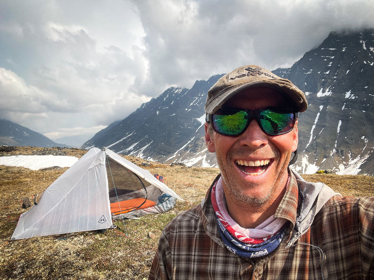 Man with sunglasses and plaid shirt in front of a tent and mountains