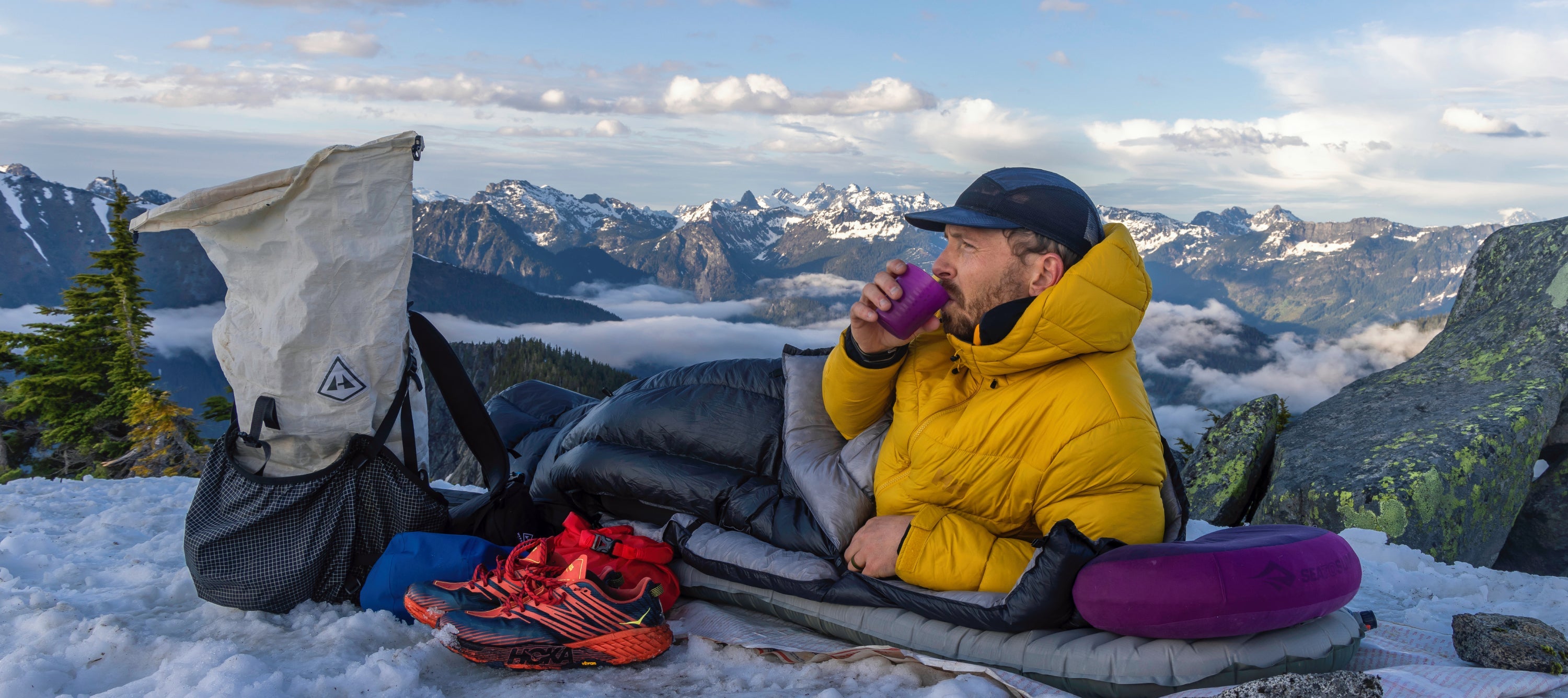 A man sitting on top of a mountain with a sleeping bag.