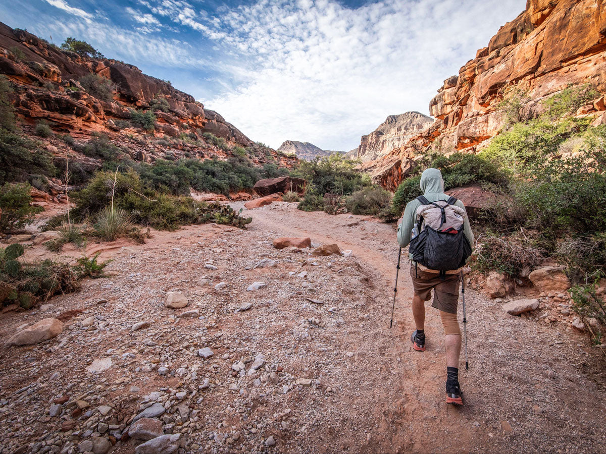 Hiker with a backpack walking on a trail in a desert landscape with rocky cliffs and sparse vegetation.